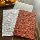 Two rectangular embossed paper sheets with a repeating fish-and-wave pattern, shown on a dark wood surface. One sheet is white and the other is terracotta brown, with a translucent sheet partially visible behind them.
