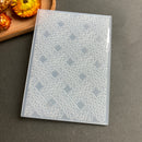 A translucent plastic sheet with an embossed geometric interlaced pattern, photographed on a dark gray surface with dried orange flowers in the upper left corner.