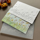 Three rectangular cards or paper pieces with embossed wildflower and grass designs are arranged on a neutral surface. One card is white with raised floral relief, one is pale green with yellow and lavender floral details, and one is translucent gray showing the negative-space embossing.
