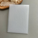 A clear plastic sheet with a repeating white chevron or herringbone pattern, photographed on a smooth gray surface with a small decorative dried-plant prop in the upper left.