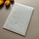 A translucent white silicone mold with embossed holiday designs of candles, mistletoe, and snowflakes, shown on a neutral gray background with dried flowers in the corner.