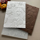Two embossed holiday-themed cards or paper sheets with pinecones, bows, and hearts, shown on a neutral surface with dried flowers in the corner. One card is white and one is brown, with a translucent silicone stamp or mold sheet visible underneath.