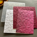 Three embossed butterfly-patterned sheets or cards in white, dusty rose, and burgundy, arranged on a gray surface with a translucent clear sheet behind them.
