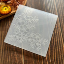 A square translucent acrylic or glass tile with an etched floral pattern, displayed diagonally on a dark wood background with dried flowers in the corner.