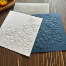 Two embossed floral paper sheets with raised cherry blossom designs, one in white and one in dark blue, displayed on a wooden surface with a translucent sheet behind them.