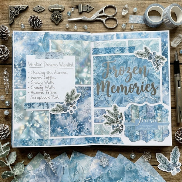 A styled flat-lay of a scrapbook journaling page with winter-themed blue and white patterned paper, silver glitter text reading 'Frozen Memories,' decorative botanical stickers, and surrounding crafting supplies on a wooden surface.