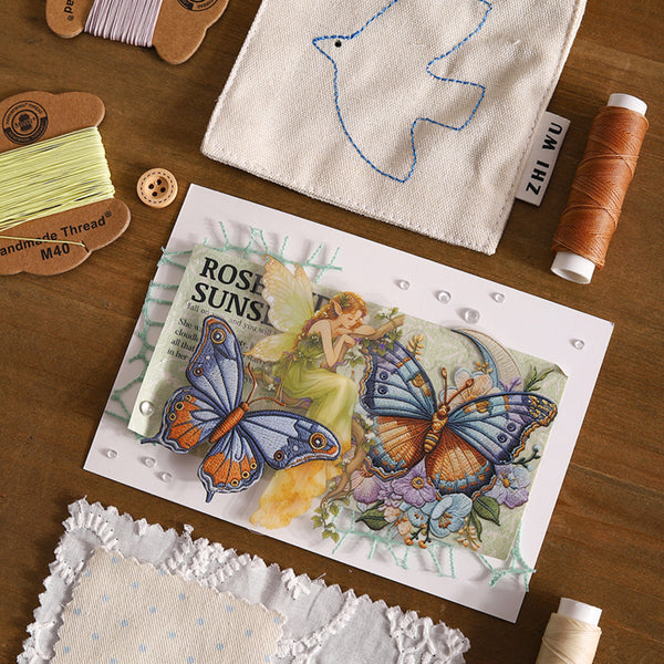 A styled flat-lay of sewing and embroidery supplies on a wooden surface, featuring a central handmade card with a fairy illustration and large blue-and-orange butterflies, surrounded by spools of thread, fabric pieces, and a white stitched bird applique.