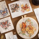 A styled flat-lay of embroidered fabric pieces featuring orange butterflies and floral designs, arranged on a wooden surface with a wooden embroidery hoop in the foreground.