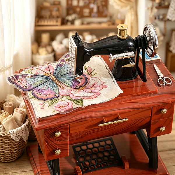 A miniature decorative sewing machine model in black with brass-colored accents, displayed on a reddish-brown wooden table. A colorful embroidered butterfly and floral fabric piece rests on top, alongside small scissors and a basket of fabric rolls, set against a softly blurred vintage-style workshop background.