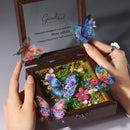 A wooden keepsake box with a floral message insert, shown open and held by two hands. The box contains colorful butterfly decorations and a lush miniature flower bed, with additional butterflies being placed on and around the box by the hands.
