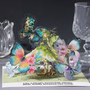 A 3D pop-up cardboard puzzle featuring a whimsical fairy surrounded by colorful butterflies and flowers, displayed on a card with text, set against a dark background with glassware.