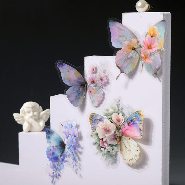Four pastel butterfly and floral wall decals arranged on white stepped display blocks against a black background. A small white cherub figurine sits on the left block.