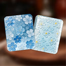 Two square decorative cards or coasters with a floral embossed pattern, one in shades of blue and white and the other in pale blue with white flowers and small yellow centers, shown against a blurred warm brown background.