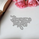 Decorative metal word die on a white surface with pink roses and an open book in the background.