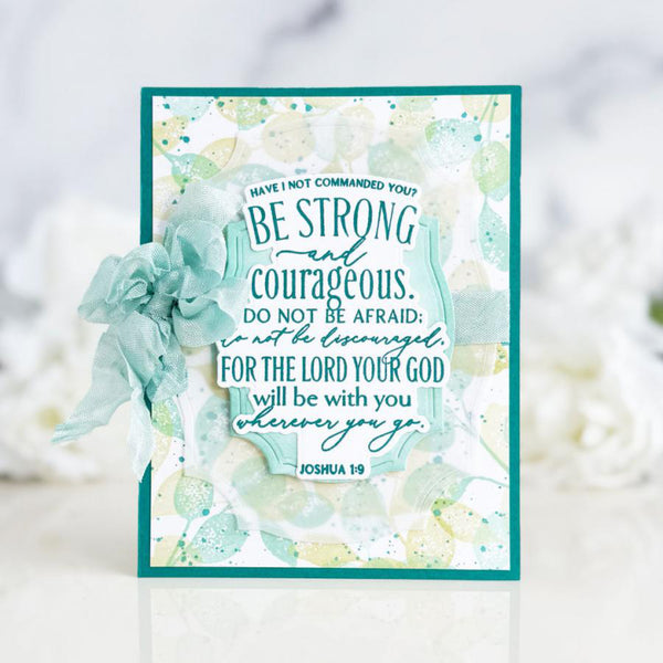 A handmade inspirational greeting card featuring a teal and white watercolor design with the Bible verse Joshua 1:9. The card is decorated with a matching teal fabric flower on the left side and is set against a soft, blurred white background.