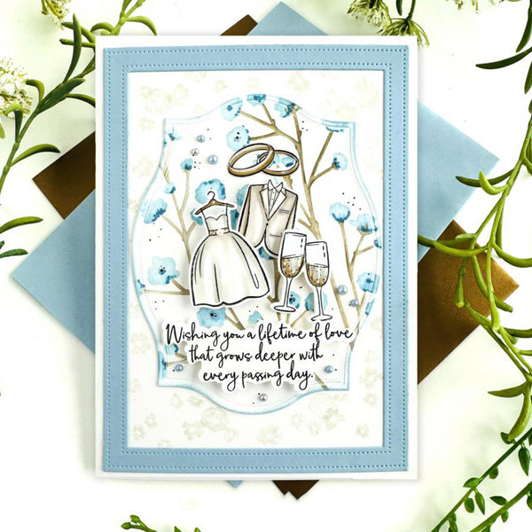 A square layered card with a light blue border and a central illustration of a wedding couple, rings, two champagne glasses, and the phrase 'Wishing you a lifetime of love that grows deeper with every passing day.' The card is styled on a white surface with blue and brown paper backing and green floral sprigs around it.