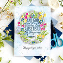 A square handmade greeting card with a floral wreath design and Bible verse text, styled in a flat lay with blue envelopes, scissors, dried flowers, and a candle on a white surface.