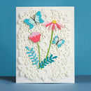 A handmade paper card featuring a white scalloped lace-like background with layered paper flowers and butterflies in pink, blue, green, and yellow, set against a solid blue background.