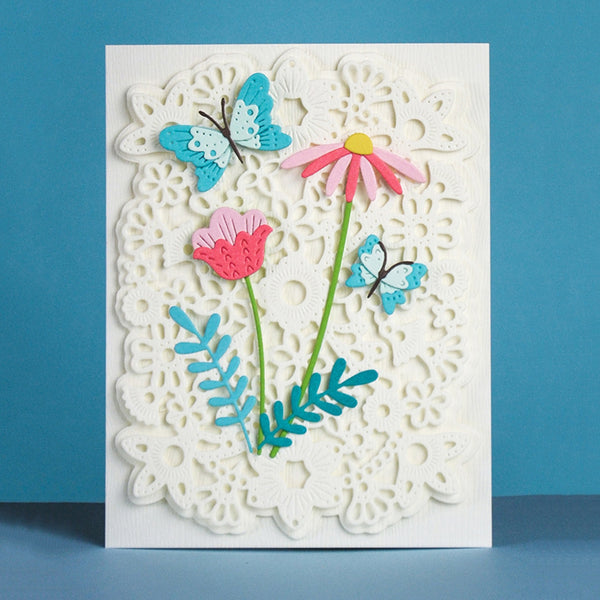 A handmade paper card featuring a white scalloped lace-like background with layered paper flowers and butterflies in pink, blue, green, and yellow, set against a solid blue background.