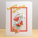 A handmade layered paper card with 'happy birthday' in yellow script, a coral scalloped border, and 3D flowers and butterflies in pink, peach, white, and green, standing on a light wood surface against a neutral gray background.