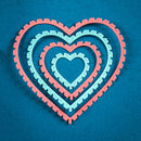 Three nested heart-shaped metal cutters or stencils in coral pink and light blue, arranged concentrically on a textured dark blue background.