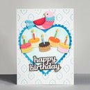 A handmade birthday card featuring a white background with light blue dotted patterns, a large light blue heart outline with scalloped blue edges, a pink and blue bird, five layered birthday cakes with candles, and the words 'happy birthday' in brown script. The card is photographed standing upright on a gray surface against a light gray background.