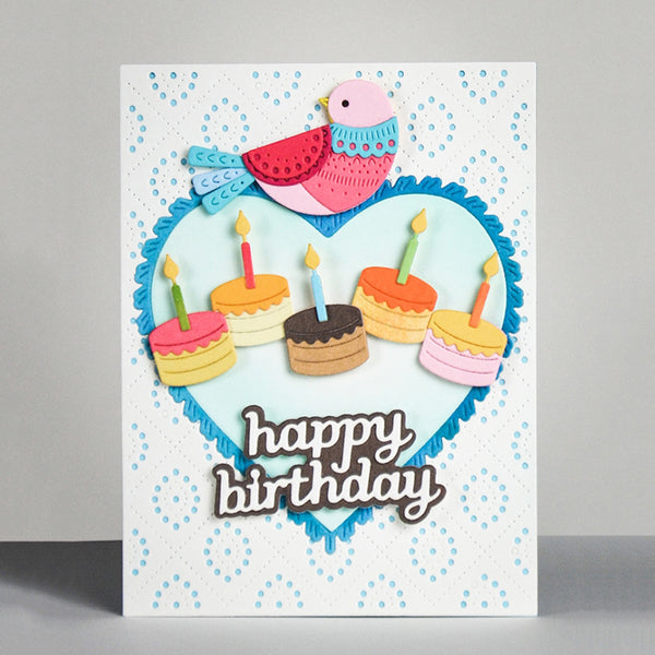A handmade birthday card featuring a white background with light blue dotted patterns, a large light blue heart outline with scalloped blue edges, a pink and blue bird, five layered birthday cakes with candles, and the words 'happy birthday' in brown script. The card is photographed standing upright on a gray surface against a light gray background.