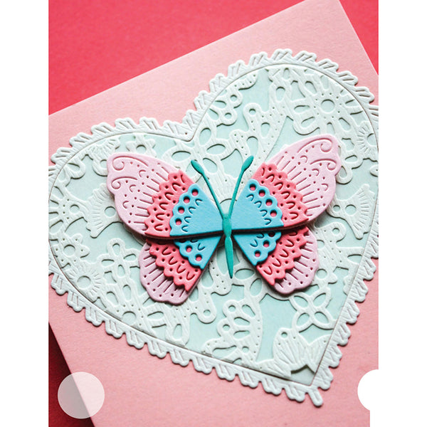 A handmade layered paper butterfly embellishment in pink and light blue, layered over a large white embossed heart-shaped lace pattern on a pink cardstock background.