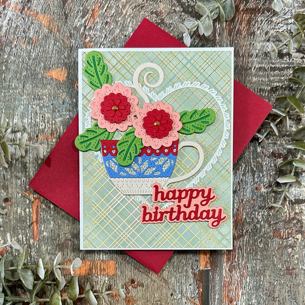 A handmade birthday card featuring a blue patterned teacup filled with pink and red flowers and green leaves, placed on top of a red envelope over a rustic wood surface with decorative greenery.