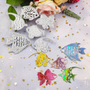 A set of fish-shaped craft cutters and decals displayed on a light pastel background with gold star confetti and floral props. The set includes several silver metal cutters and multiple colorful iridescent decals in blue, yellow, pink, purple, green, and orange.