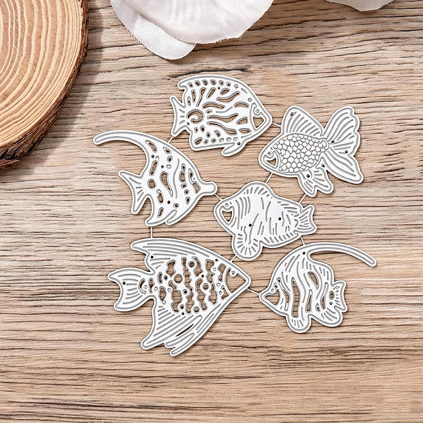 Six white laser-cut metal fish dies with detailed tropical fish patterns, arranged on a light brown wooden surface.