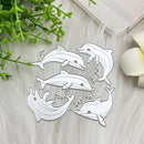 A set of five white die-cut dolphin shapes with silver metal outlines, arranged on a white faux fur surface with decorative green leaves and a white flower.