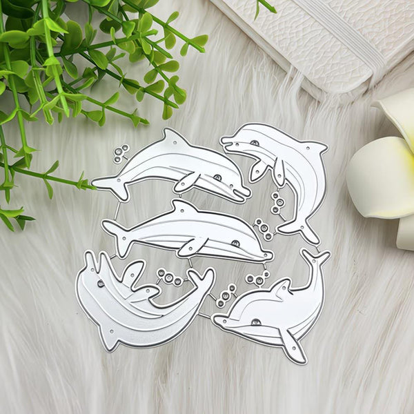 A set of five white die-cut dolphin shapes with silver metal outlines, arranged on a white faux fur surface with decorative green leaves and a white flower.