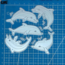 A set of five dolphin-shaped metal cutting dies arranged on a blue gridded craft cutting mat. The dies are silver-colored metal with pre-cut holes, and the mat features measurement markings.
