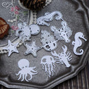 A set of white metal cutters shaped like sea creatures and nautical motifs, including octopus, starfish, crab, seahorse, jellyfish, and turtle, arranged on a textured gray fabric with decorative props.