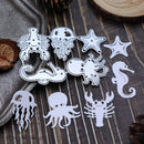 A set of white metal cutout templates featuring various sea life designs including an octopus, seahorse, starfish, jellyfish, and crab, arranged on a dark wood background with decorative props.