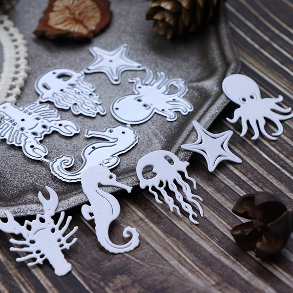 White metal nail art dies shaped like sea creatures, including a seahorse, octopus, starfish, jellyfish, and lobster, arranged on a gray textured cloth over a dark wood background with decorative dried botanicals.