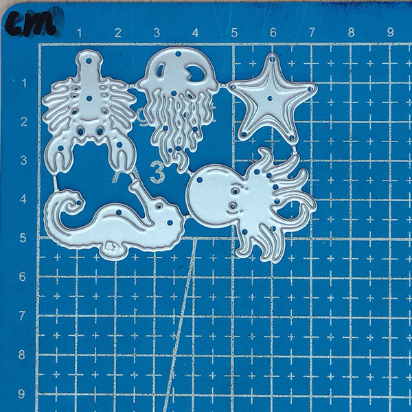 A set of five silver metal die-cut craft shapes, including a starfish, octopus, seahorse, jellyfish, and turtle, arranged on a blue grid cutting mat.
