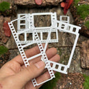 A hand holds two white plastic miniature film strip frames in front of a miniature wooden tabletop scene with small green moss and brown bark details.