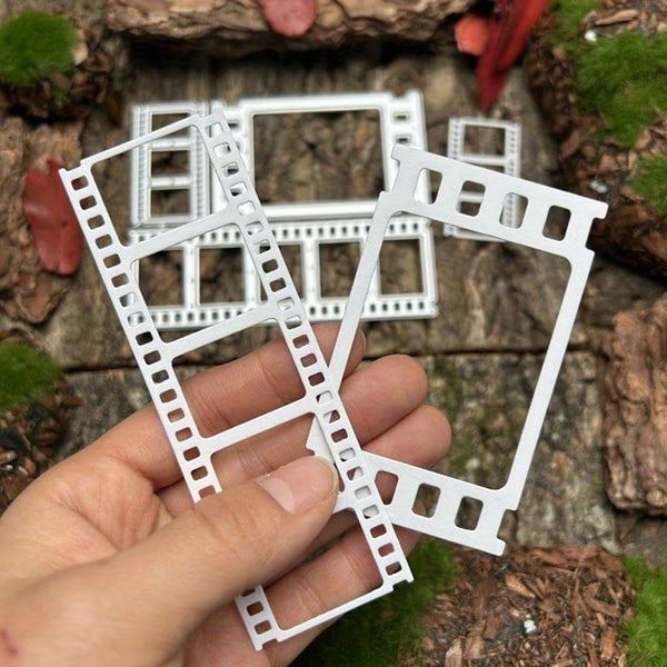 A hand holds two white plastic miniature film strip frames in front of a miniature wooden tabletop scene with small green moss and brown bark details.
