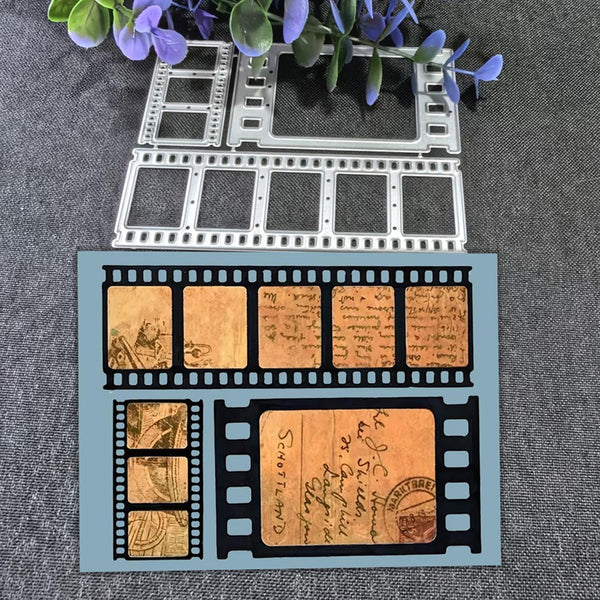 A flat sheet of cardstock or craft paper featuring a vintage film strip design with antique lettering and sepia-toned document illustrations, displayed on a gray textured fabric background with decorative purple flowers and metal film-frame cutters above it.