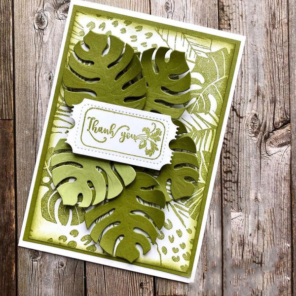 A handmade tropical-themed greeting card featuring layered olive green monstera leaf cutouts over a white and light green patterned background, with a white scalloped label reading 'thank you' and a small floral design, displayed on a rustic wooden surface.