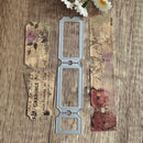 A metal die-cut stamping template with three rectangular slots is displayed on a rustic wooden surface, flanked by decorative scrapbook paper strips featuring vintage French text and floral patterns, with small white flowers at the top.