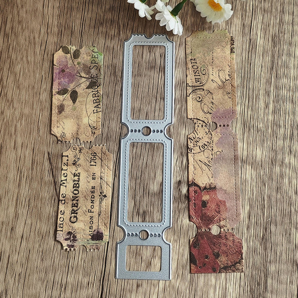 A metal die-cut stamping template with three rectangular slots is displayed on a rustic wooden surface, flanked by decorative scrapbook paper strips featuring vintage French text and floral patterns, with small white flowers at the top.