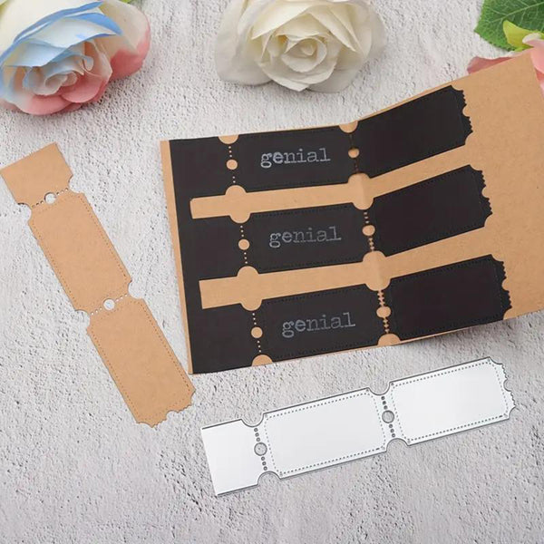 A craft kit featuring a black and kraft paper envelope with the word 'genial' printed on the inside, accompanied by separate blank ticket-shaped pieces and a metal scoring ruler, arranged on a textured white surface with decorative paper flowers.