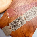 Two vintage-style Christmas movie ticket ornaments displayed on a wooden surface. One ticket is a dusty rose pink, and the other is a textured beige/tan color.