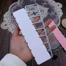 A hand holds a white paper template with a silver metal die-cutting frame over a dark wood surface with a lace mat and pink paper visible in the background.