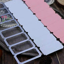 A close-up of a craft die set with rectangular scoring and perforation dies in silver metal, shown alongside pink and white paper sheets on a dark wooden surface.