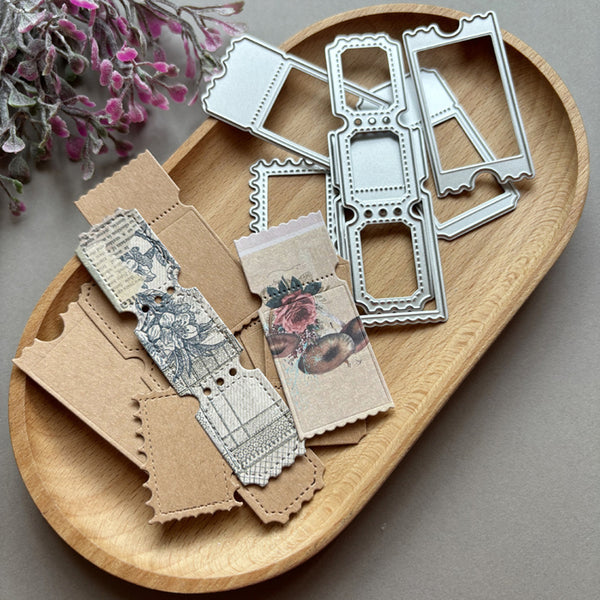 A set of metal embossing dies with vintage-style cutout frames and patterned paper sheets arranged on a round wooden tray, with dried flowers in the top-left corner.