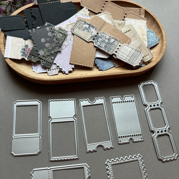A set of silver metal embossing dies displayed on a gray surface, with a wooden tray holding matching decorative paper scraps above them.